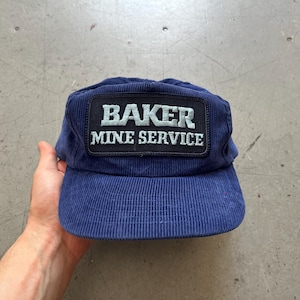 May include: A navy blue corduroy cap featuring a rectangular patch with the words "BAKER MINE SERVICE" in white. The cap has a structured peak and a classic, casual design.