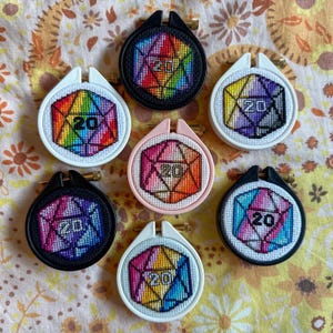 Può includere: Una collezione di sette piccoli ricami a punto croce rotondi. Ogni pezzo presenta un dado D20 geometrico e colorato con il numero 20. Le cornici sono nere, bianche e rosa. Lo sfondo ha un motivo floreale.