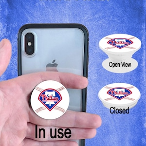 Könnte beinhalten: Ein weißer Handy-Popsocket mit einem roten, weißen und blauen Baseball-Design. Das Design zeigt das Wort "Phillies" in blauer Schrift. Der Popsocket ist sowohl in geöffneter als auch in geschlossener Position dargestellt.