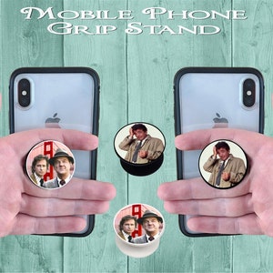 Könnte beinhalten: Handy-Halterung mit Pop-out-Design. Der Ständer hat eine schwarze Basis und ein rundes Bild einer Fernsehsendung. Der Text "Mobile Phone Grip Stand" befindet sich oben im Bild.