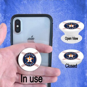 May include: A white and blue Houston Astros phone popper in use, with an open and closed view. The popper features the team logo on a baseball design. The text "PHONE POPPER" and "In use" are also visible.