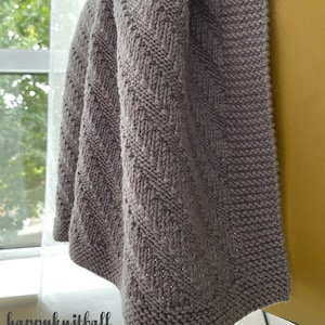 May include: A gray knitted baby blanket with a chevron pattern. The blanket is draped over a chair.