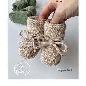 May include: A pair of beige knitted baby booties with bow ties, held by a hand. The booties are made of a soft, textured yarn. The text "The Little Wuggies" is at the top, and "PDF Pattern" is in a circle.