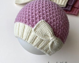 May include: A purple and white knitted hat with a bow on top. The hat is made with a textured stitch and has a soft, cozy feel.