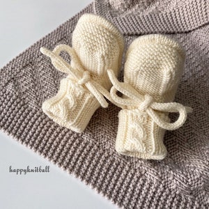 May include: A pair of cream-colored knitted baby booties featuring a cable knit pattern on the cuffs and matching tie closures. The booties are placed on a textured, light brown knitted fabric with the text happyknitball printed in black.