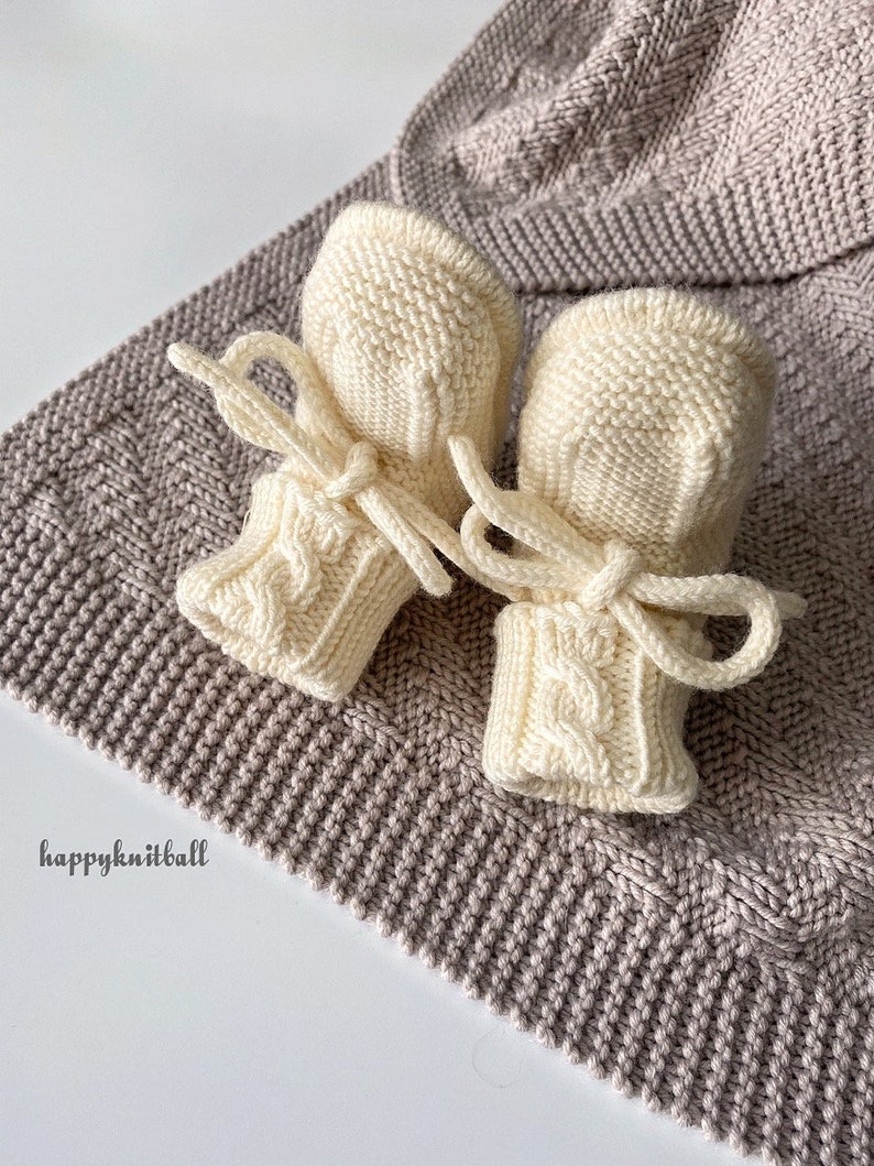 May include: A pair of cream-colored knitted baby booties featuring a cable knit pattern on the cuffs and matching tie closures. The booties are placed on a textured, light brown knitted fabric with the text happyknitball printed in black.