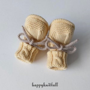 May include: A pair of pale yellow knitted baby mittens featuring a cable knit pattern on the cuffs and a smooth knit texture on the hand section. Each mitten is tied with a light gray cord. The text "happyknitball" is printed below the mittens.