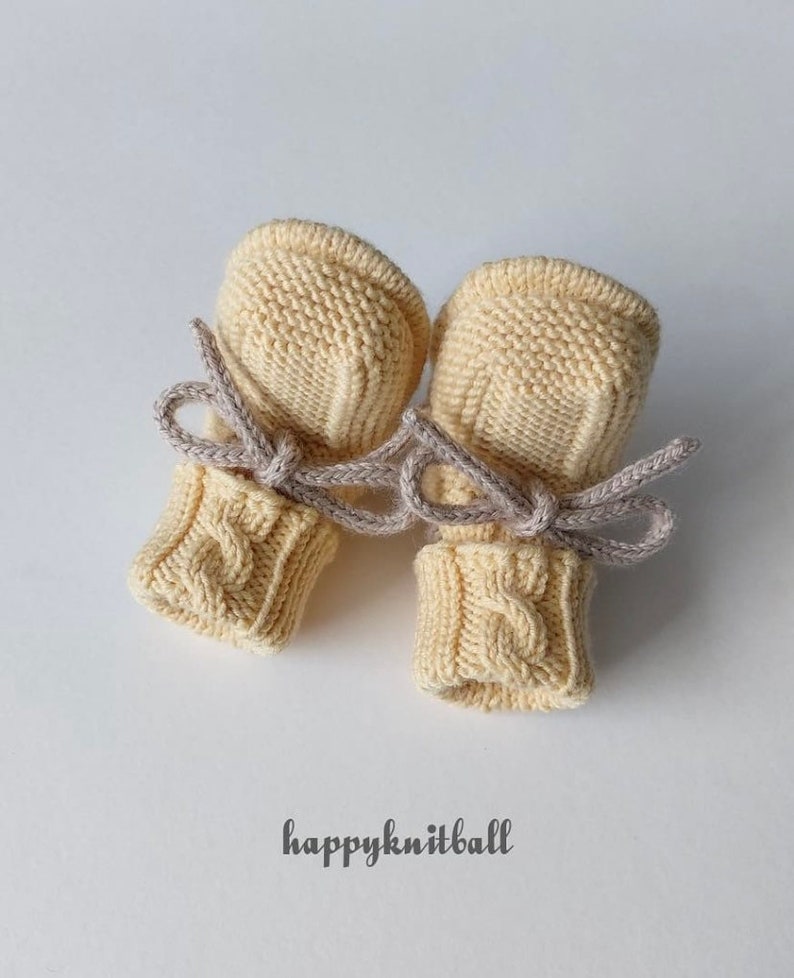 May include: A pair of pale yellow knitted baby mittens featuring a cable knit pattern on the cuffs and a smooth knit texture on the hand section. Each mitten is tied with a light gray cord. The text "happyknitball" is printed below the mittens.