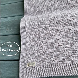 May include: A gray knitted blanket with a subtle textured pattern. The blanket is made with a soft yarn and has a finished edge. The label "happyknitball" is visible on the blanket.