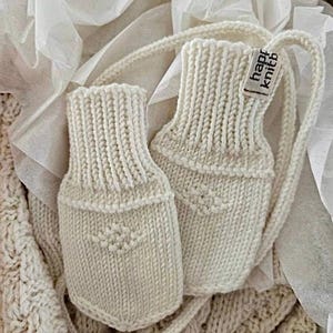 May include: A pair of off-white knitted mittens featuring a ribbed cuff and a small diamond-shaped textured pattern on the palm. A small fabric tag attached to the side of one mitten displays the text "happy knitb" in black lettering.