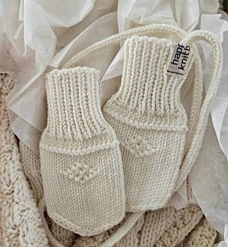 May include: A pair of off-white knitted mittens featuring a ribbed cuff and a small diamond-shaped textured pattern on the palm. A small fabric tag attached to the side of one mitten displays the text "happy knitb" in black lettering.