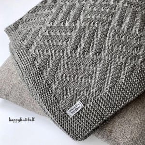 May include: A grey knitted blanket featuring a textured herringbone pattern and a garter stitch border. A small white fabric label with the text "happyknitball" is sewn onto the corner. The word "happyknitball" is also printed in black.