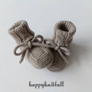May include: A pair of light brown knitted booties featuring a cable knit pattern on the cuffs and a tied drawstring closure at the ankle. The text "happyknitball" is printed in a dark grey script font below the footwear on a plain background.
