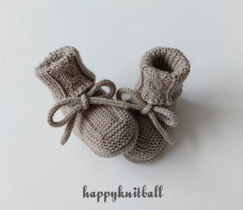May include: A pair of light brown knitted booties featuring a cable knit pattern on the cuffs and a tied drawstring closure at the ankle. The text "happyknitball" is printed in a dark grey script font below the footwear on a plain background.