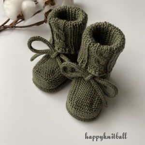 May include: A pair of olive green knitted booties featuring a ribbed cuff with a cable knit pattern and a matching tie closure at the ankle. The text happyknitball is printed in the lower right corner of the image near the knitted footwear.