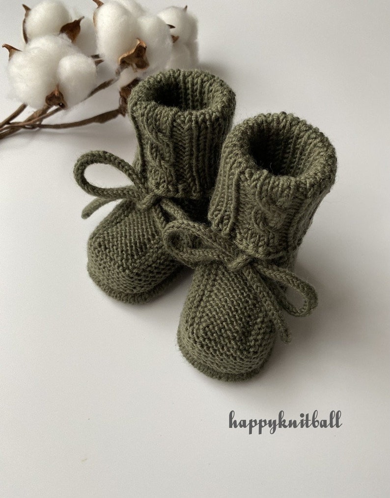 May include: A pair of olive green knitted booties featuring a ribbed cuff with a cable knit pattern and a matching tie closure at the ankle. The text happyknitball is printed in the lower right corner of the image near the knitted footwear.