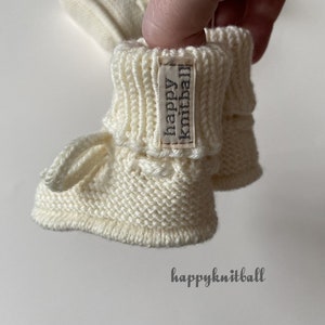 May include: A pair of off-white knitted baby booties featuring a ribbed cuff and a small fabric tag with the text happy knitball. The booties have a textured garter stitch pattern on the foot and a small loop detail. The text happyknitball is visible.