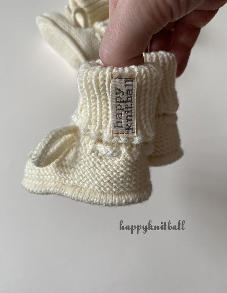 May include: A pair of off-white knitted baby booties featuring a ribbed cuff and a small fabric tag with the text happy knitball. The booties have a textured garter stitch pattern on the foot and a small loop detail. The text happyknitball is visible.