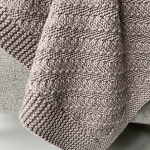 May include: A close-up of a soft, knitted blanket in a light brown color. The blanket has a chevron pattern and is folded over on itself. The blanket is sitting on a light gray surface.