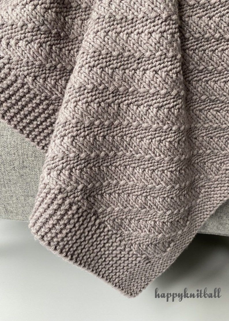 May include: A close-up of a soft, knitted blanket in a light brown color. The blanket has a chevron pattern and is folded over on itself. The blanket is sitting on a light gray surface.