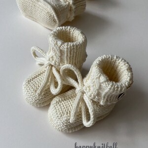 May include: A pair of cream-colored knitted baby booties with ribbed cuffs and matching tie closures, accompanied by a second pair in the background. The text "happyknitball" is printed in the lower right corner of the image.