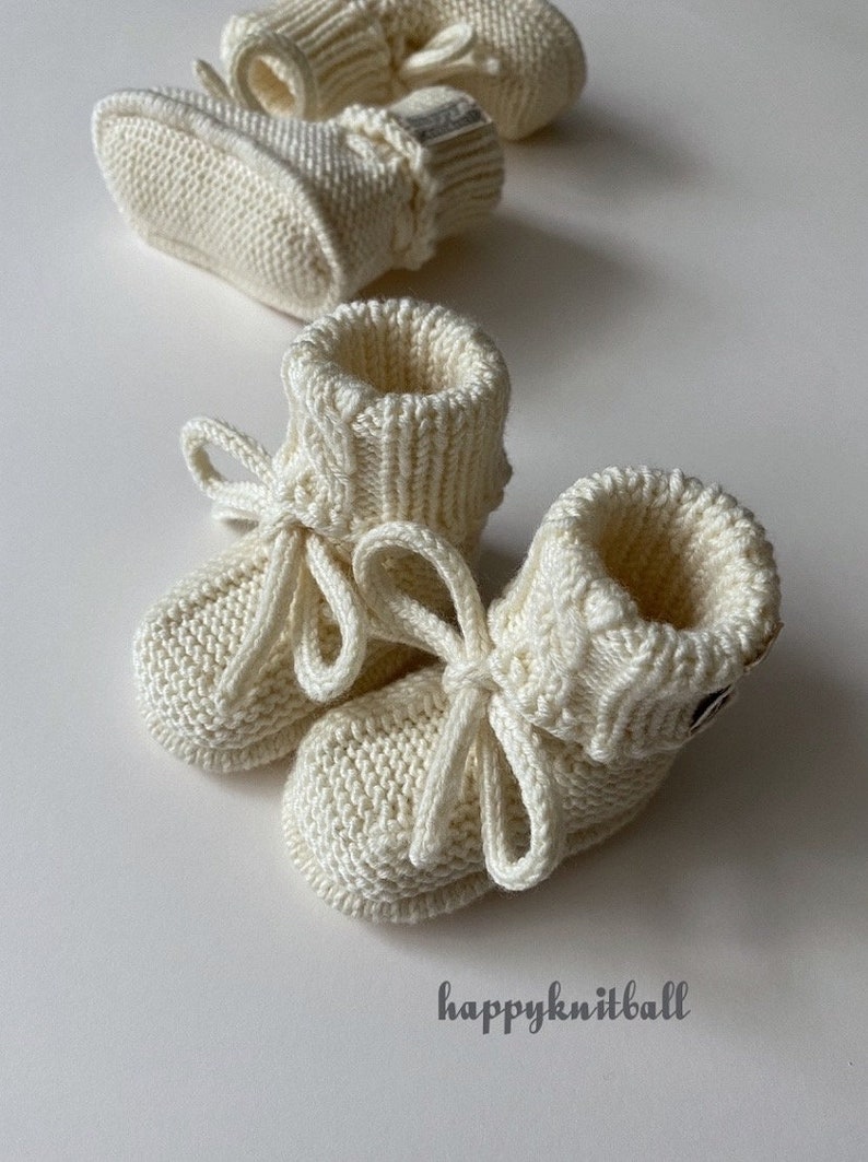 May include: A pair of cream-colored knitted baby booties with ribbed cuffs and matching tie closures, accompanied by a second pair in the background. The text "happyknitball" is printed in the lower right corner of the image.