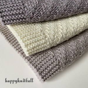 May include: Three knitted blankets stacked on top of each other. The top and bottom blankets are a light gray color, and the middle blanket is a cream color. The blankets are made with a textured stitch.
