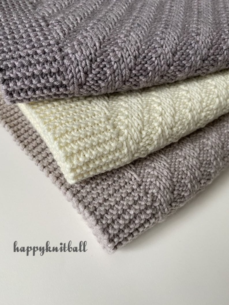 May include: Three knitted blankets stacked on top of each other. The top and bottom blankets are a light gray color, and the middle blanket is a cream color. The blankets are made with a textured stitch.
