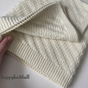 May include: A white knitted blanket with a chevron pattern. The blanket is folded in half and held by a hand. The text "happyknitball" is visible in the lower left corner of the image.