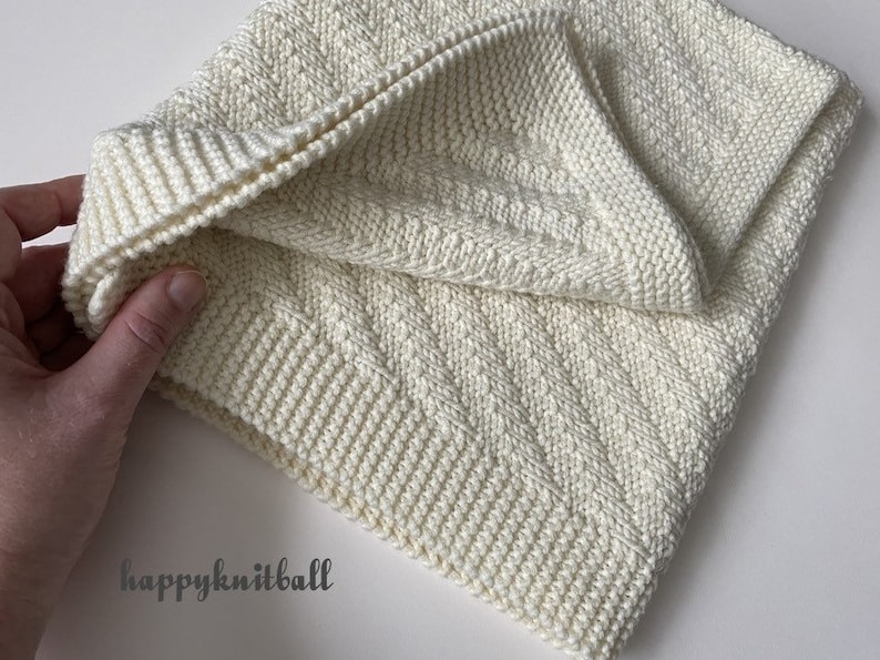 May include: A white knitted blanket with a chevron pattern. The blanket is folded in half and held by a hand. The text "happyknitball" is visible in the lower left corner of the image.