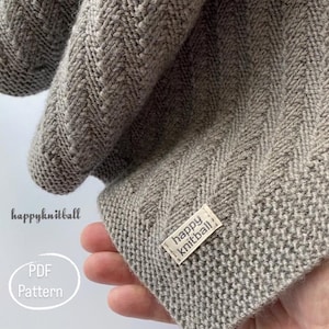 Könnte beinhalten: Nahaufnahme eines grauen Strickstoffs mit Fischgrätmuster. Der Stoff ist gefaltet und zeigt die Textur und die Stichdetails. Ein kleines Etikett mit dem Text "happy knitball" ist angebracht.