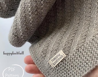 Könnte beinhalten: Nahaufnahme eines grauen Strickstoffs mit Fischgrätmuster. Der Stoff ist gefaltet und zeigt die Textur und die Stichdetails. Ein kleines Etikett mit dem Text "happy knitball" ist angebracht.