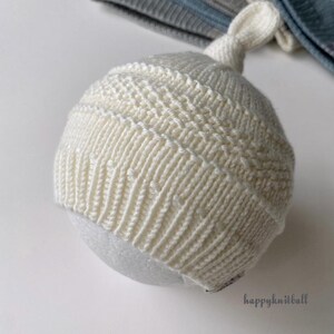 May include: A cream-colored knit hat with a top knot, featuring a ribbed brim and a textured horizontal band pattern. The hat is displayed on a round form, with additional folded knit hats in blue and gray tones visible in the background.