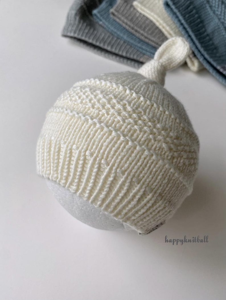May include: A cream-colored knit hat with a top knot, featuring a ribbed brim and a textured horizontal band pattern. The hat is displayed on a round form, with additional folded knit hats in blue and gray tones visible in the background.