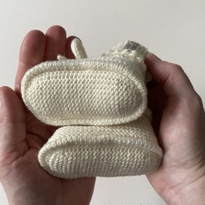 May include: A pair of cream-colored hand-knitted booties featuring a textured stitch pattern and a small fabric tag on the cuff. The booties are stacked vertically, showing the oval-shaped soles. The text happyknitball is visible below.