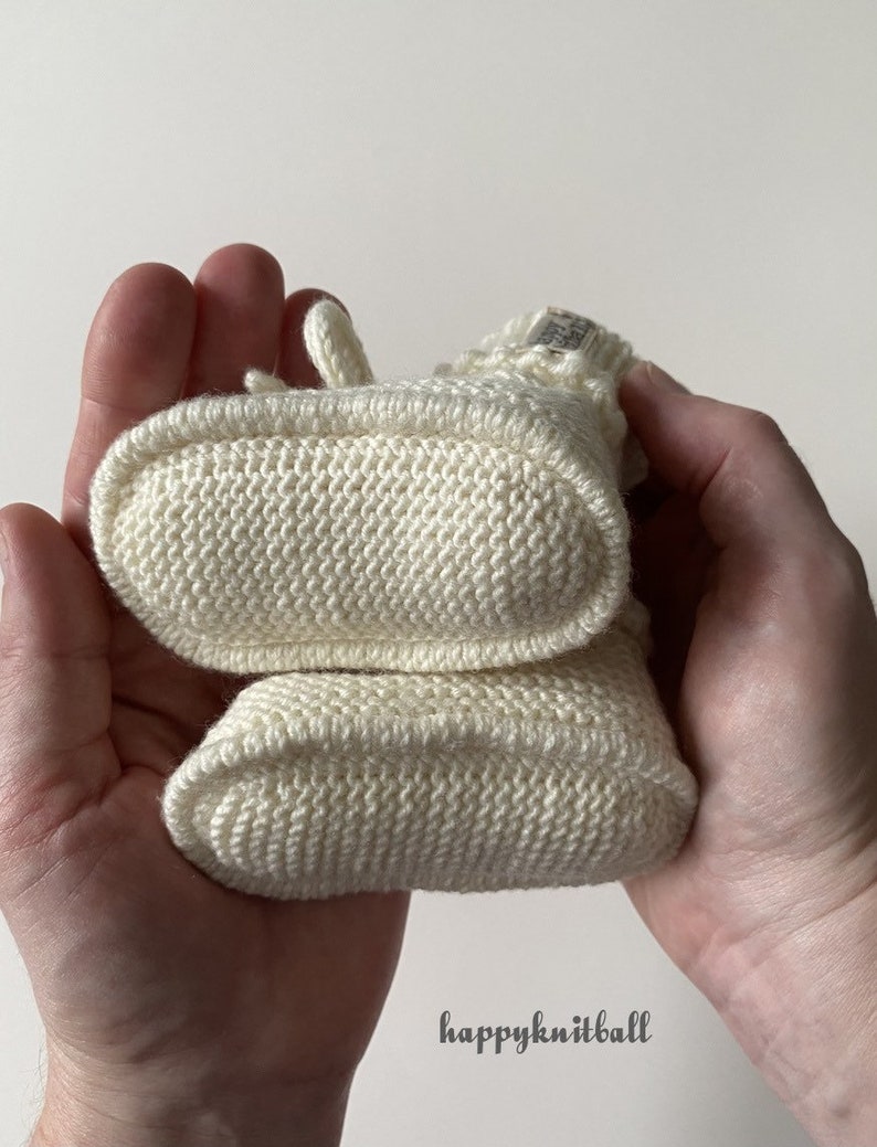 May include: A pair of cream-colored hand-knitted booties featuring a textured stitch pattern and a small fabric tag on the cuff. The booties are stacked vertically, showing the oval-shaped soles. The text happyknitball is visible below.