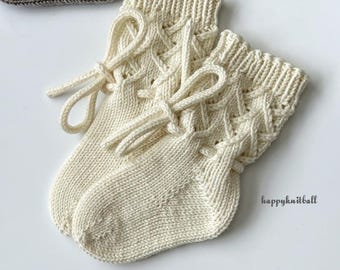 May include: A pair of cream-colored knitted socks featuring a ribbed cuff, a lattice cable pattern, and a decorative drawstring tie. The socks are crafted with a smooth knit foot section and include the text happyknitball.