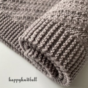May include: A close-up of a folded knitted blanket in a light brown color. The blanket has a textured pattern and is made with a soft yarn.