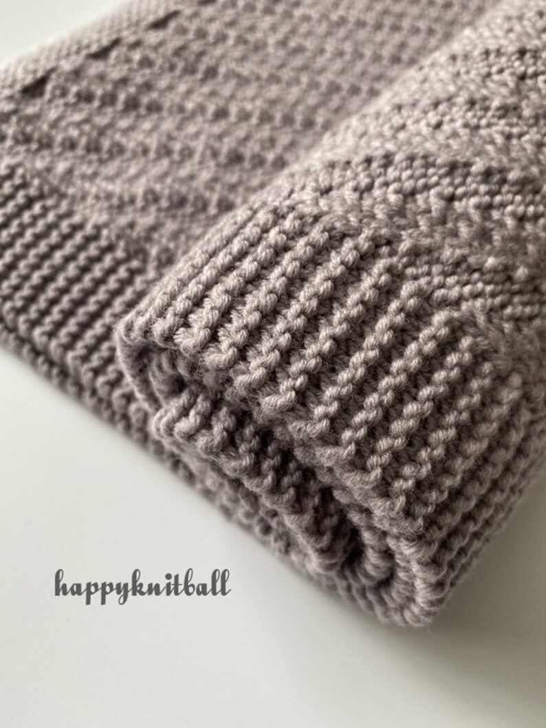 May include: A close-up of a folded knitted blanket in a light brown color. The blanket has a textured pattern and is made with a soft yarn.