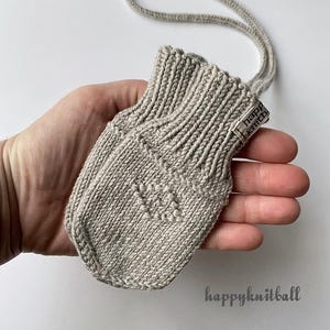 May include: A light beige knitted mitten with a ribbed cuff and a small tag reading "happy knitball" attached to the side. The mitten features a subtle textured pattern on the front and is connected to a matching knitted cord for hanging.