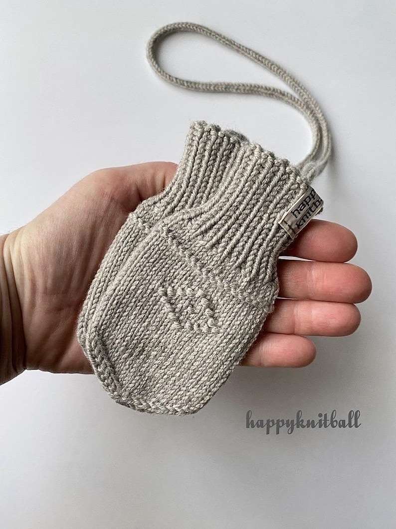 May include: A light beige knitted mitten with a ribbed cuff and a small tag reading "happy knitball" attached to the side. The mitten features a subtle textured pattern on the front and is connected to a matching knitted cord for hanging.