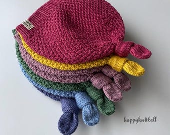 May include: A stack of five knitted hats in magenta, yellow, mauve, green, and blue, each featuring a knotted top detail. A small beige fabric tag with the text happyknitball is attached to the magenta hat, and the text happyknitball is printed in black on the right.