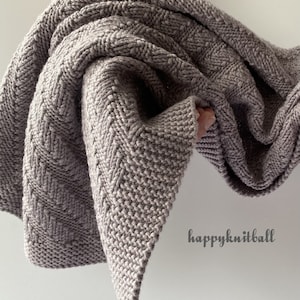May include: A soft, gray knitted blanket with a chevron pattern. The blanket is folded and draped over a hand. The text "happyknitball" is visible in the lower right corner of the image.