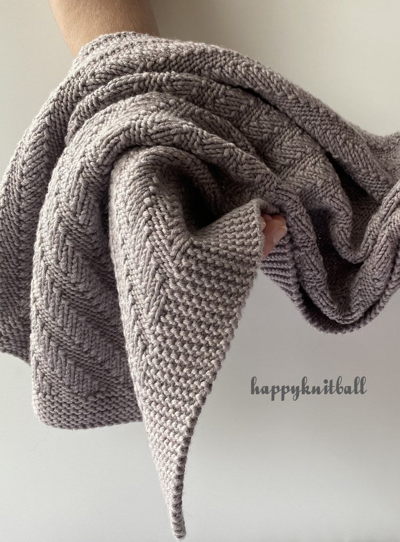 May include: A soft, gray knitted blanket with a chevron pattern. The blanket is folded and draped over a hand. The text "happyknitball" is visible in the lower right corner of the image.