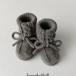 May include: A pair of charcoal gray knitted booties featuring a ribbed cuff, a textured garter stitch foot, and a matching knitted drawstring tie tied in a bow at the front. The text "happyknitball" is printed in black below the booties.