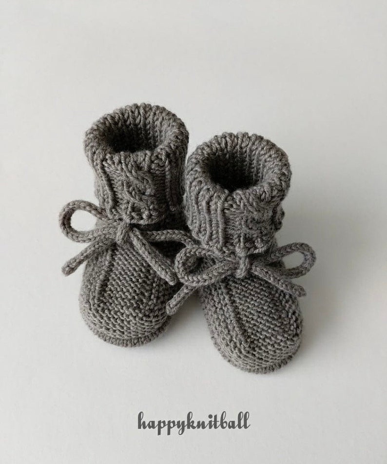 May include: A pair of charcoal gray knitted booties featuring a ribbed cuff, a textured garter stitch foot, and a matching knitted drawstring tie tied in a bow at the front. The text "happyknitball" is printed in black below the booties.
