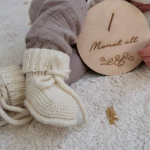May include: A pair of cream-colored knitted baby booties with ribbed cuffs and tie closures, paired with a round wooden milestone disc engraved with the number 1 and the German text Monat alt, featuring a small decorative branch illustration.