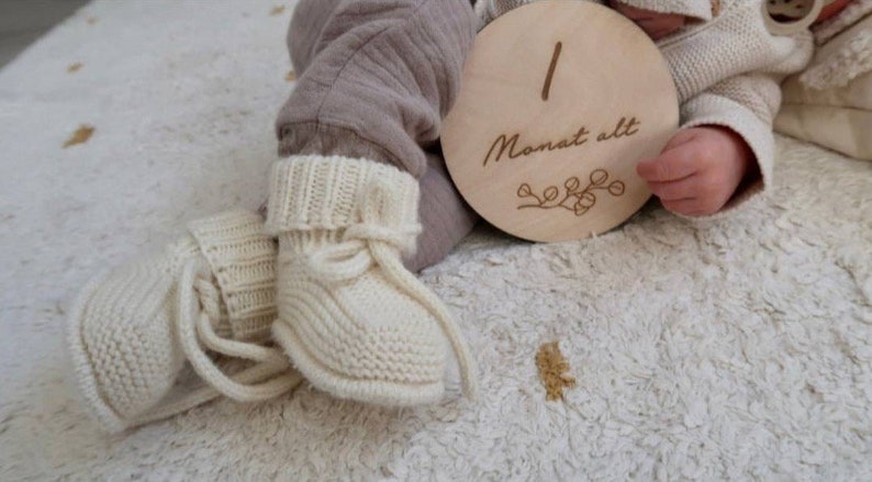 May include: A pair of cream-colored knitted baby booties with ribbed cuffs and tie closures, paired with a round wooden milestone disc engraved with the number 1 and the German text Monat alt, featuring a small decorative branch illustration.
