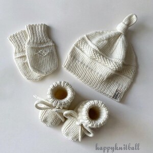 May include: A set of off-white knitted baby accessories including a pair of mittens, a knotted hat, and booties with tie closures. The hat features a small rectangular tag with the text "happy knitball" and the same text appears below.