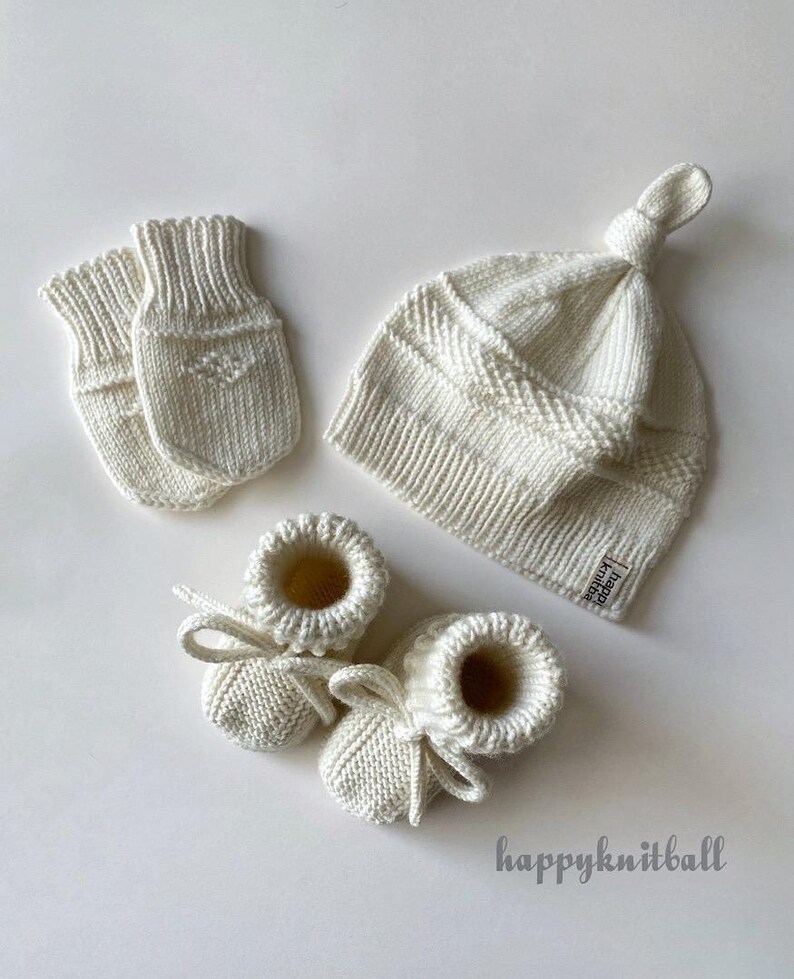 May include: A set of off-white knitted baby accessories including a pair of mittens, a knotted hat, and booties with tie closures. The hat features a small rectangular tag with the text "happy knitball" and the same text appears below.